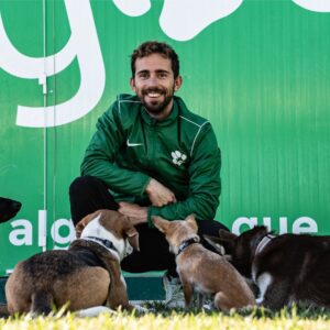 Entrenador sonriente de Argos Adiestramiento Canino rodeado de perros variados, sentado frente a un fondo verde con el logo de Argos