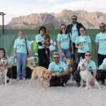 Miembros del Club de Argos y sus perros posando frente a banners promocionales en una competición al aire libre en las instalaciones de Argos con montañas en el fondo